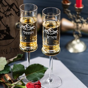 May include: Two clear glass champagne flutes with gold rims. The flutes are engraved with mountain silhouettes and the names "Markus" and "Jenni".