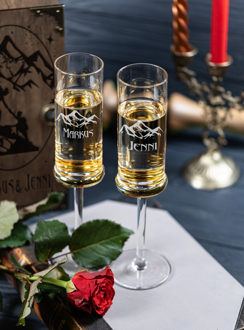 May include: Two clear glass champagne flutes with gold rims. The flutes are engraved with mountain silhouettes and the names "Markus" and "Jenni".
