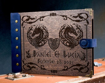 May include: A wooden wedding album with a dark finish, featuring engraved dragon designs and the names "DANIEL & LUCIA" with the date "September 28, 2023." The album has a blue leather spine and a matching clasp.