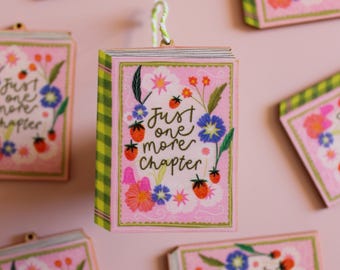 May include: Pink, floral-themed book-shaped ornament with the text "Just one more chapter." The cover features strawberries, blue and orange flowers, and green leaves. The spine has a green and yellow checkered pattern. A white and green string is attached for hanging.