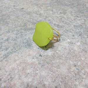 Può includere: Un anello verde lime con una pietra quadrata con angoli arrotondati. La pietra è attaccata a una fascia di filo dorato. L'anello è appoggiato su una superficie screziata grigio chiaro. Gioielli.