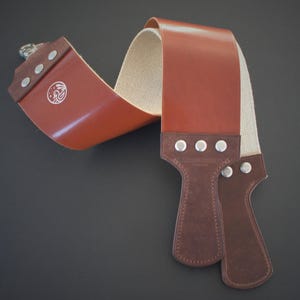 May include: A brown leather strop with a tan canvas backing, used for sharpening straight razors. The strop features a brown leather handle with rivets and a decorative emblem. The leather is a rich, reddish-brown color.