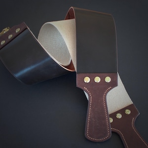 May include: A leather strop for sharpening straight razors. The strop features a dark brown leather exterior, a tan canvas interior, and a dark brown handle with brass rivets. The handle is stitched with white thread.