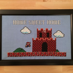 May include: A framed cross-stitch design featuring a pixelated image of a video game level with a castle, clouds, and a character jumping. The text "Home Sweet Home" is stitched above the level.