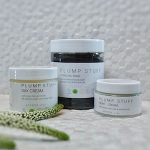 May include: Three white jars of skin care products with black lids. The jars are labeled "Plump Stuff" and contain a day cream, charcoal mask, and night cream. The jars are sitting on a white tiled surface with white flowers in the foreground.