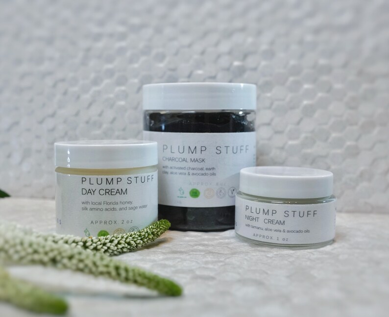 May include: Three white jars of skin care products with black lids. The jars are labeled "Plump Stuff" and contain a day cream, charcoal mask, and night cream. The jars are sitting on a white tiled surface with white flowers in the foreground.
