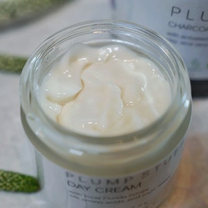 May include: A white jar of Plum Petuff Day Cream with a white lid. The cream is visible through the glass jar. The jar is labeled with the text "Plum Petuff Day Cream" and "with local Florida honey, silk amino acids, and a...