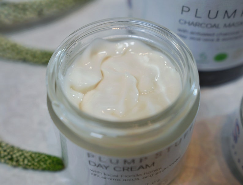 May include: A white jar of Plum Petuff Day Cream with a white lid. The cream is visible through the glass jar. The jar is labeled with the text "Plum Petuff Day Cream" and "with local Florida honey, silk amino acids, and a...