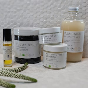 May include: A set of four skincare products in white jars and bottles with black labels. The products include a charcoal mask, a night cream, a day cream, and a facial toner. The products are labeled "Plump Stuff" and feature ingredients like charcoal, aloe vera, avocado oil, and Florida honey.