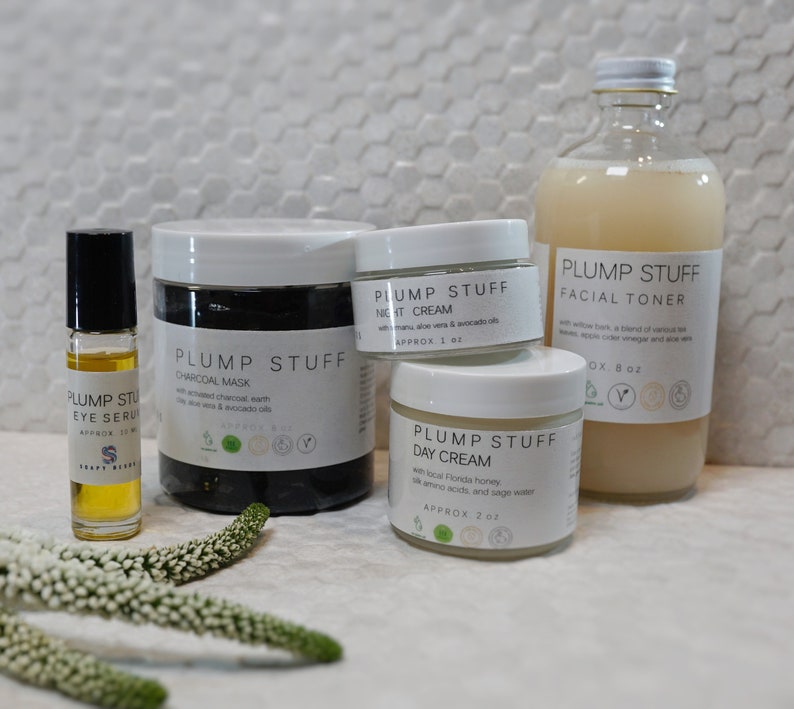 May include: A set of four skincare products in white jars and bottles with black labels. The products include a charcoal mask, a night cream, a day cream, and a facial toner. The products are labeled "Plump Stuff" and feature ingredients like charcoal, aloe vera, avocado oil, and Florida honey.