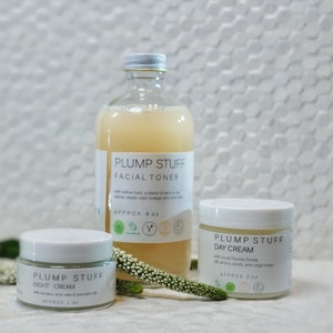 May include: Three glass bottles of skincare products with white labels. The bottles are labeled "Plump Stuff" and contain facial toner, day cream, and night cream. The bottles are on a white tiled surface with a small green plant.