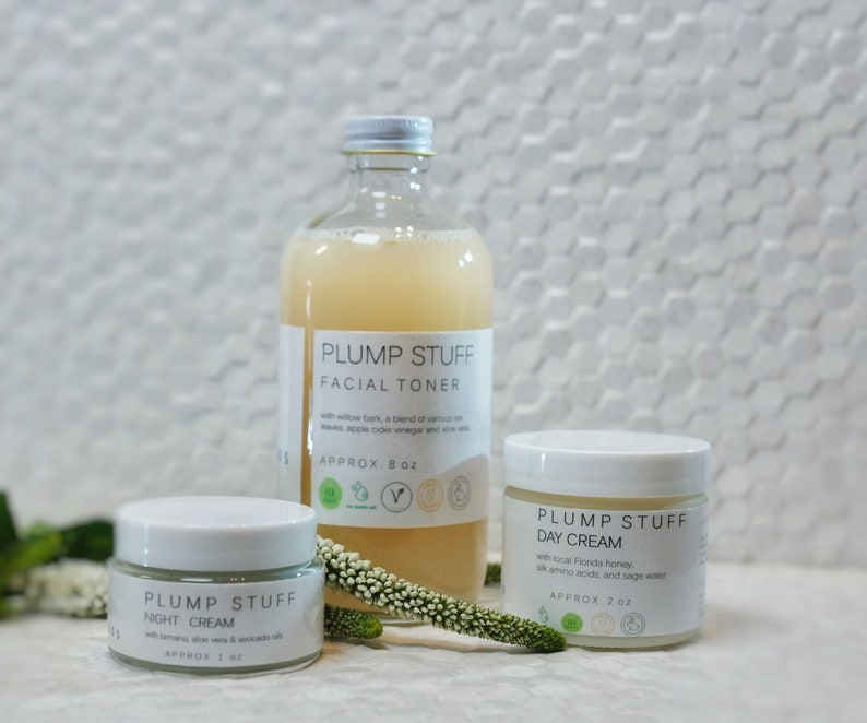 May include: Three glass bottles of skincare products with white labels. The bottles are labeled "Plump Stuff" and contain facial toner, day cream, and night cream. The bottles are on a white tiled surface with a small green plant.