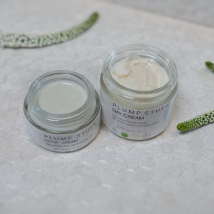 May include: Two glass jars of face cream with white labels. The jar on the left says "PLUMP STUFF NIGHT CREAM" and the jar on the right says "PLUMP STUFF DAY CREAM". Both jars have a white cream inside.