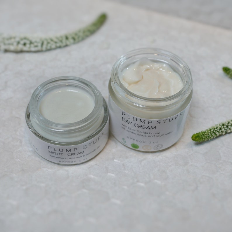 May include: Two glass jars of face cream with white labels. The jar on the left says "PLUMP STUFF NIGHT CREAM" and the jar on the right says "PLUMP STUFF DAY CREAM". Both jars have a white cream inside.