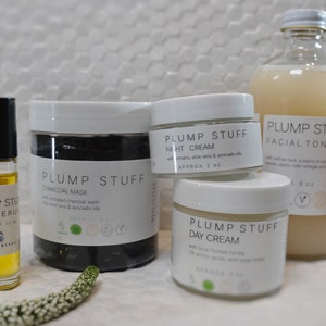 May include: A set of four skincare products in white jars and a glass bottle. The products are labeled "Plump Stuff" and include a charcoal mask, night cream, day cream, and facial toner. The products are all natural and organic.