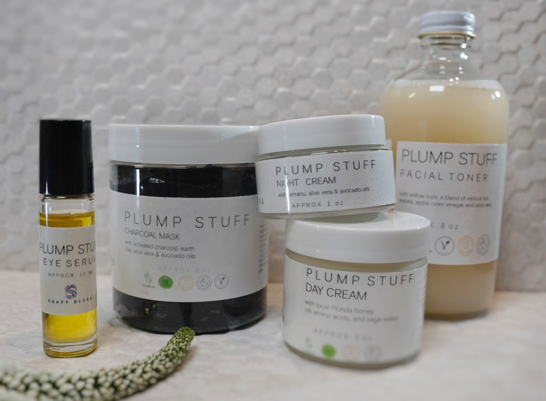 May include: A set of four skincare products in white jars and a glass bottle. The products are labeled "Plump Stuff" and include a charcoal mask, night cream, day cream, and facial toner. The products are all natural and organic.