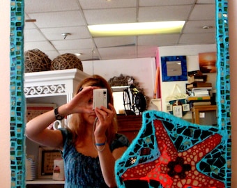Op de afbeelding: Een turquoise mozaïek ingelijste spiegel met een rood en zwart mozaïek zeester ontwerp in de rechter benedenhoek. Een vrouw wordt in de spiegel gereflecteerd terwijl ze een selfie maakt.