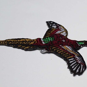 May include: An embroidered patch of a pheasant in flight. The pheasant is brown, red, green, and white with black outlines. The patch is perfect for adding a touch of nature to your clothing or accessories.