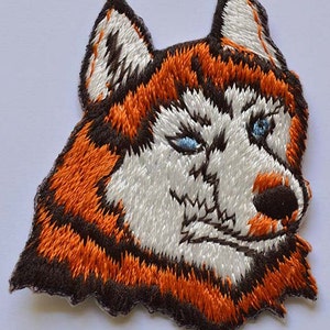 May include: Embroidered patch of a husky dog's face with brown, orange, white, and blue fur. The dog is winking with one eye.