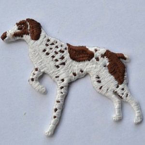 May include: White and brown embroidered iron-on patch of a dog. The dog is standing with its legs spread apart and its head turned to the left. The dog has brown spots on its body.