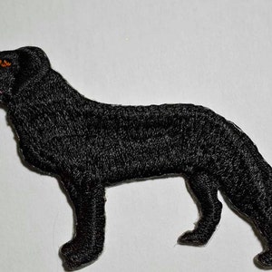 May include: Embroidered black dog patch. The dog is depicted in a side profile, with its mouth slightly open, revealing a pink tongue. The eyes are orange. The patch is made of a textured, black thread and is likely designed to be sewn onto clothing or accessories.