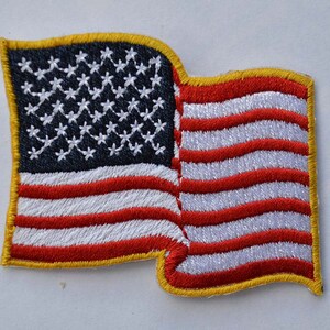 May include: A red, white, and blue embroidered American flag patch with a gold border. The flag is waving in the wind.