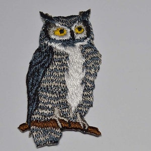 May include: Embroidered iron-on patch of a gray and white owl with yellow eyes perched on a brown branch.