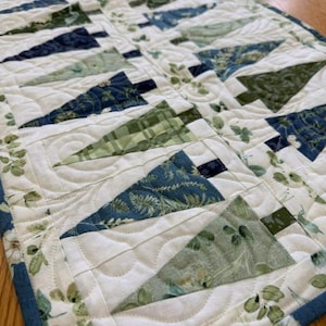 May include: A quilted table runner featuring a repeating pattern of triangular tree shapes in various shades of blue and green. The fabric blocks are set against a white background with a floral print, finished with intricate swirl stitching.