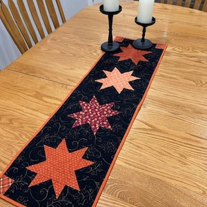 May include: A rectangular fabric table runner featuring a black background with a gold-colored leaf pattern and four appliqued stars in varying shades of orange, red, and plaid. The runner has a solid orange border and square corner accents.