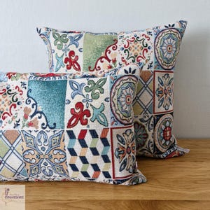 May include: Two decorative throw pillows with a patchwork design. The pillows feature a variety of colorful patterns, including floral, geometric, and mosaic-style tiles in shades of blue, red, green, and cream. The pillows are placed on a wooden surface.