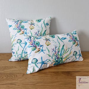 May include: Two white rectangular throw pillows with a watercolor design of sea life. The pattern includes blue starfish, coral, seashells, and seaweed in shades of blue, green, and orange. The pillows are on a wooden surface.