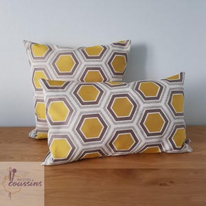 Può includere: Due cuscini rettangolari decorativi con un motivo geometrico a nido d'ape. Il motivo presenta esagoni gialli, grigi e marroni su sfondo bianco. I cuscini sono appoggiati su una superficie di legno.