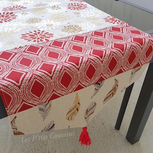 Può includere: Runner da tavolo decorativo con vari disegni. Presenta motivi geometrici rossi e bianchi, fiori beige e piume. Un bordo rosso e un fiocco rosso adornano le estremità. Il testo "Les P'tits Coussins" è visibile.