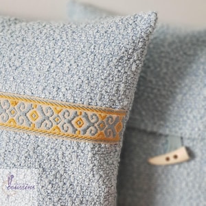 Könnte beinhalten: Nahaufnahme von zwei hellblauen, strukturierten Zierkissen. Ein Kissen hat ein dekoratives Band mit einem gelben und grauen geometrischen Muster. Das andere Kissen hat einen Knopfverschluss mit einem weißen Knopf.