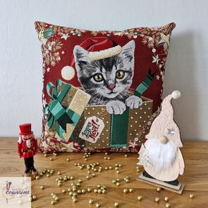 May include: A square throw pillow featuring a woven design of a gray tabby kitten wearing a red Santa hat, peeking out of a gold gift box with a green bow and a tag that reads Joyeux Noël, set against a dark red background with pine branches.