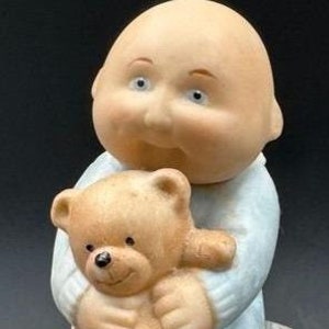 May include: A porcelain figurine of a bald baby holding a brown teddy bear. The baby is wearing a light blue onesie.