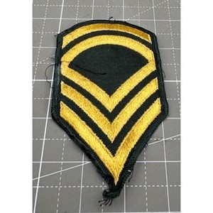 May include: A black and gold military rank patch with three gold stripes. The patch is shaped like a chevron and has a black background.