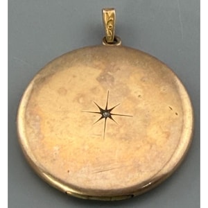 May include: A round, gold-colored locket with a starburst design and a small, central gem. The locket has a decorative bail for a chain. The surface shows slight wear, suggesting it is vintage.