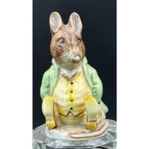 May include: A brown and white ceramic figurine of a mouse wearing a green jacket and yellow waistcoat. The mouse is sitting with its legs crossed and has a small tail.