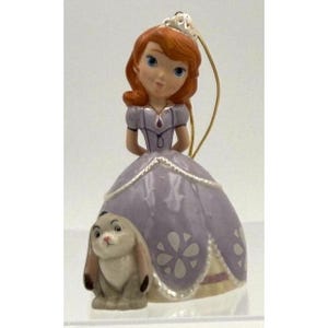 May include: A figurine of a princess with auburn hair, wearing a lavender gown with floral accents. A small rabbit is at the base of the dress. The figurine has a gold string for hanging.