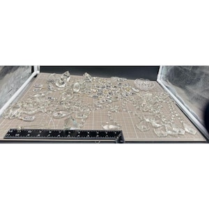 Op de afbeelding: Een verzameling heldere, facetgeslepen kristallen kralen en decoratieve elementen, waaronder druppels, ovalen en andere vormen, zijn gerangschikt op een snijmat met raster. Een zwarte liniaal met centimeter markeringen staat op de voorgrond.