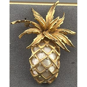 Puede incluir: Un broche de piña en tono dorado con una parte superior de hojas de metal texturizado y un cuerpo con patrón de celosía. El cuerpo de la piña está incrustado con un material blanco iridiscente y acentuado con pequeñas piedras redondas y claras.