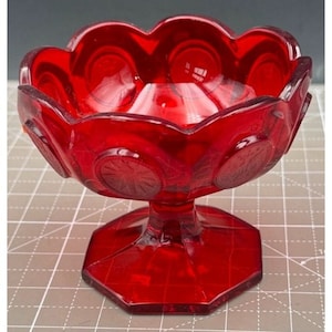 Puede incluir: Un plato de dulces de vidrio rojo con un borde festoneado y una base de pedestal. El plato tiene un patrón de pequeños círculos.