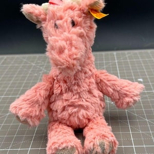 May include: A pink plush giraffe toy with a pink bow and a Steiff button tag. The giraffe is sitting on a gray and white grid background.