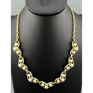 Puede incluir: Un collar dorado con una serie de bucles entrelazados, cada uno con tres perlas de imitación. El collar se exhibe en un maniquí negro. La cadena tiene un diseño de eslabones delicados y el estilo es vintage.