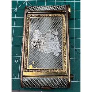 May include: A vintage silver and gold compact with a black and white patterned design. The compact features a map of Germany with the words "French Zone" and "US Zone" printed on it.