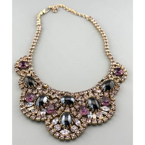 以下が含まれることがあります： ビンテージのゴールドトーンネックレス。ビブスタイルのペンダントが付いています。ペンダントには、黒、紫、クリアクリスタルの宝石が飾られています。ネックレスはニュートラルグレーの背景に表示されています。