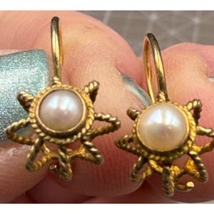May include: A pair of gold-colored earrings. Each earring features a central, round, off-white pearl surrounded by a gold-toned, starburst-shaped frame. The frame has a twisted rope design. The earrings are on a light-colored background.
