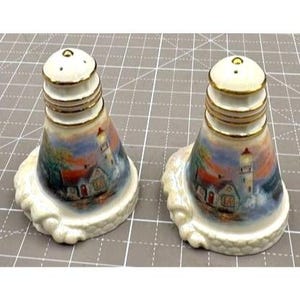Può includere: Un paio di saliere e pepiere in ceramica bianca a forma di fari con finiture dorate e una scena dipinta di un faro al tramonto.