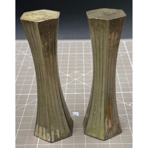 May include: Two vintage brass salt and pepper shakers. Each shaker has a hexagonal top and base, with a fluted, hourglass-shaped body. The brass has a tarnished, aged appearance, with vertical lines adding texture. The shakers are displayed on a grid surface.
